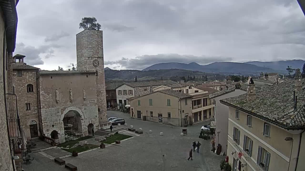 A town square with a brick building and clock tower, featuring a tree on its top, overlooks a hilly landscape under an overcast sky.