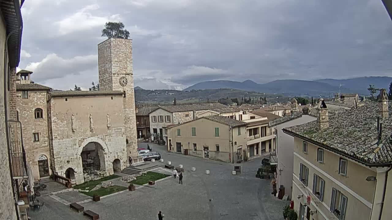 A town square with stone buildings, a clock tower topped with a tree, and distant mountains under a cloudy sky.
