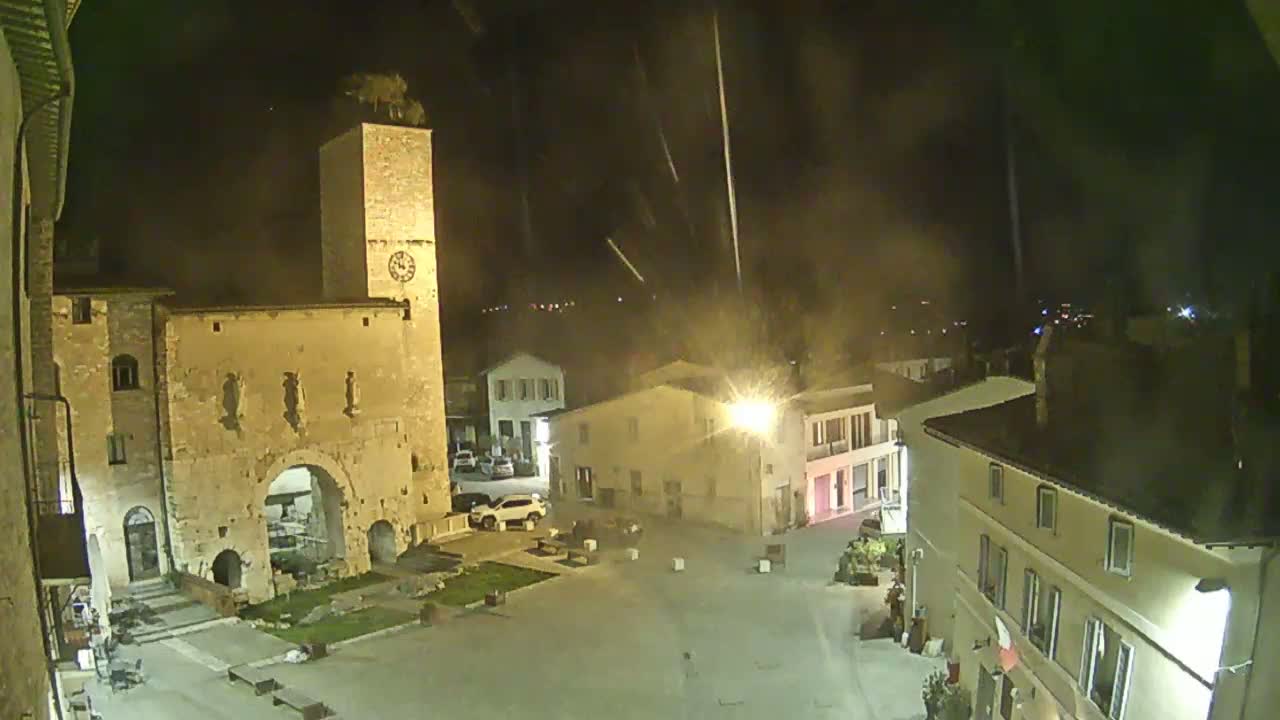 A nighttime view of a town square with ancient stone buildings, a clock tower, and a few cars, under a slightly hazy sky with streaks of light possibly from rain or fireworks.