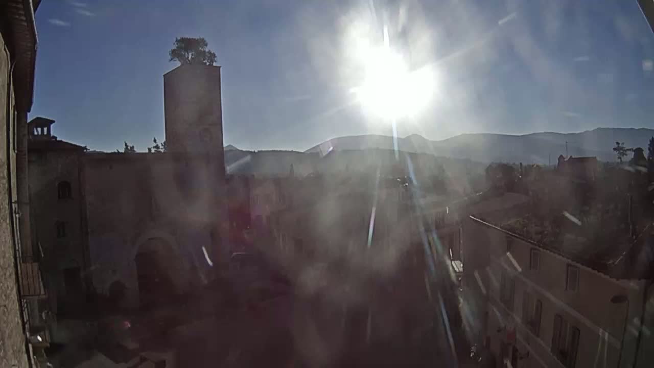 A hazy sun shines brightly over a town's stone buildings and a tall tower topped with a small tree, with distant mountains visible in the background.