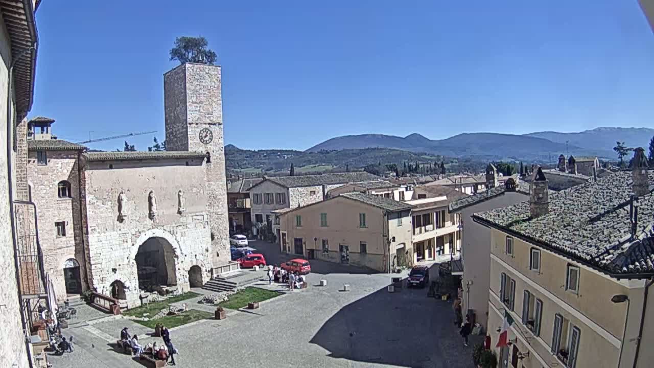 A town square with a clock tower, several buildings, and a few cars and people is visible under a clear blue sky, with distant mountains in the background.