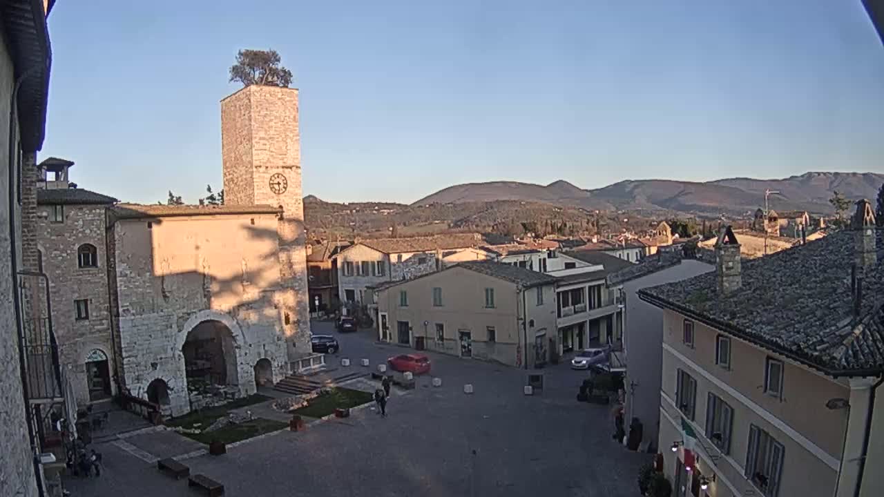 A town square with stone buildings, a clock tower topped with a tree, and distant mountains under a clear, sunny sky.