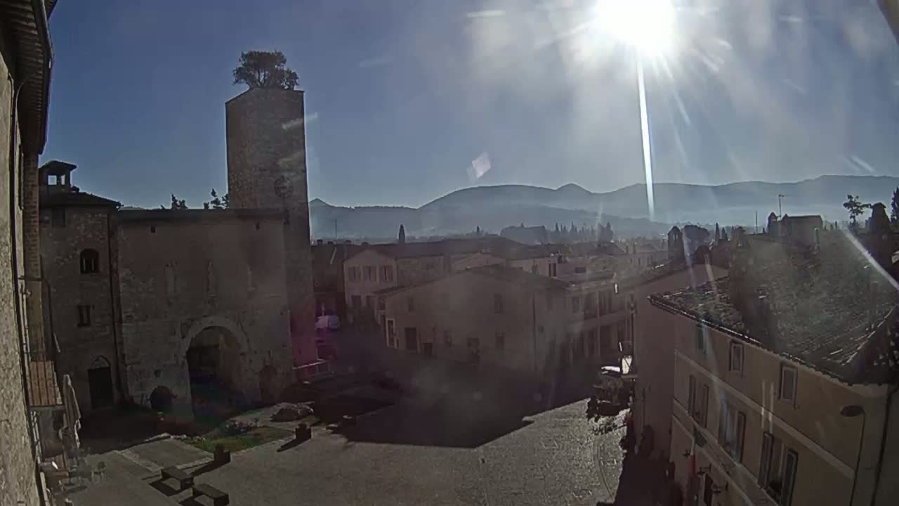 A sunny day reveals a town square surrounded by aged stone buildings, a tall tower topped with a tree, and hazy mountains in the distance.