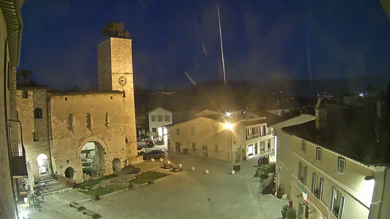 A nighttime view of a town square, illuminated by streetlights, featuring a tall stone tower with a clock, and several buildings under a clear, dark sky with faint streaks.