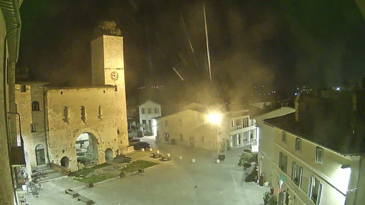 A nighttime view of a town square with ancient stone buildings, a clock tower, and light rain or mist falling.