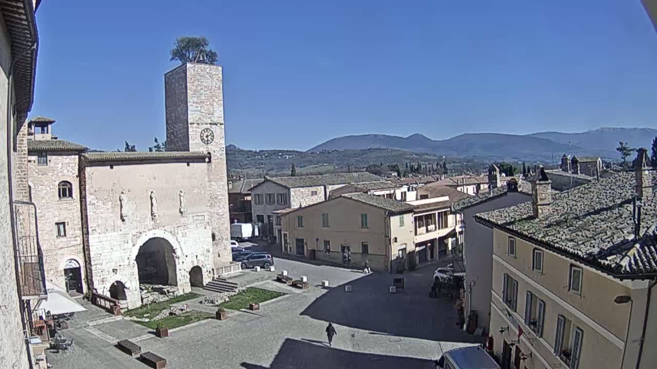 A town square with a tall clock tower and surrounding buildings is visible under a clear blue sky, with rolling hills in the background.