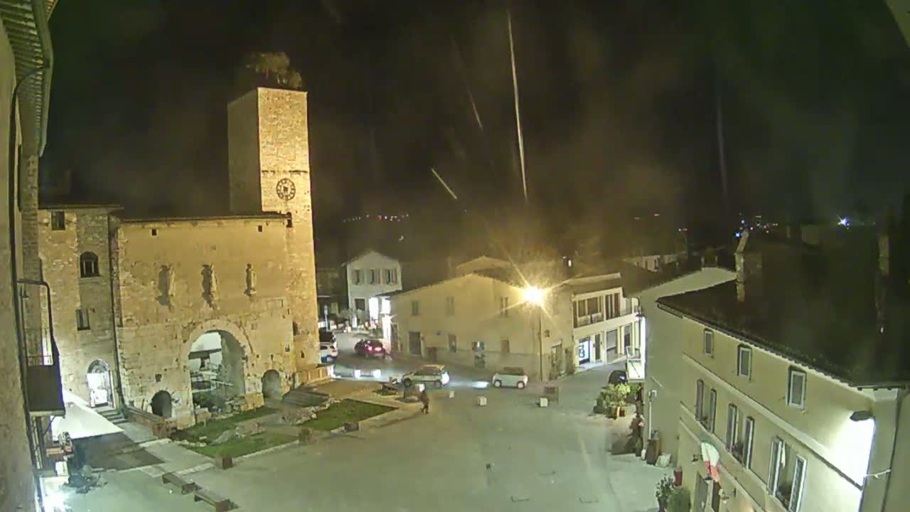 A nighttime view of a town square with several buildings, including a tall stone tower, under a slightly overcast sky with streaks of light possibly from rain or fireworks.
