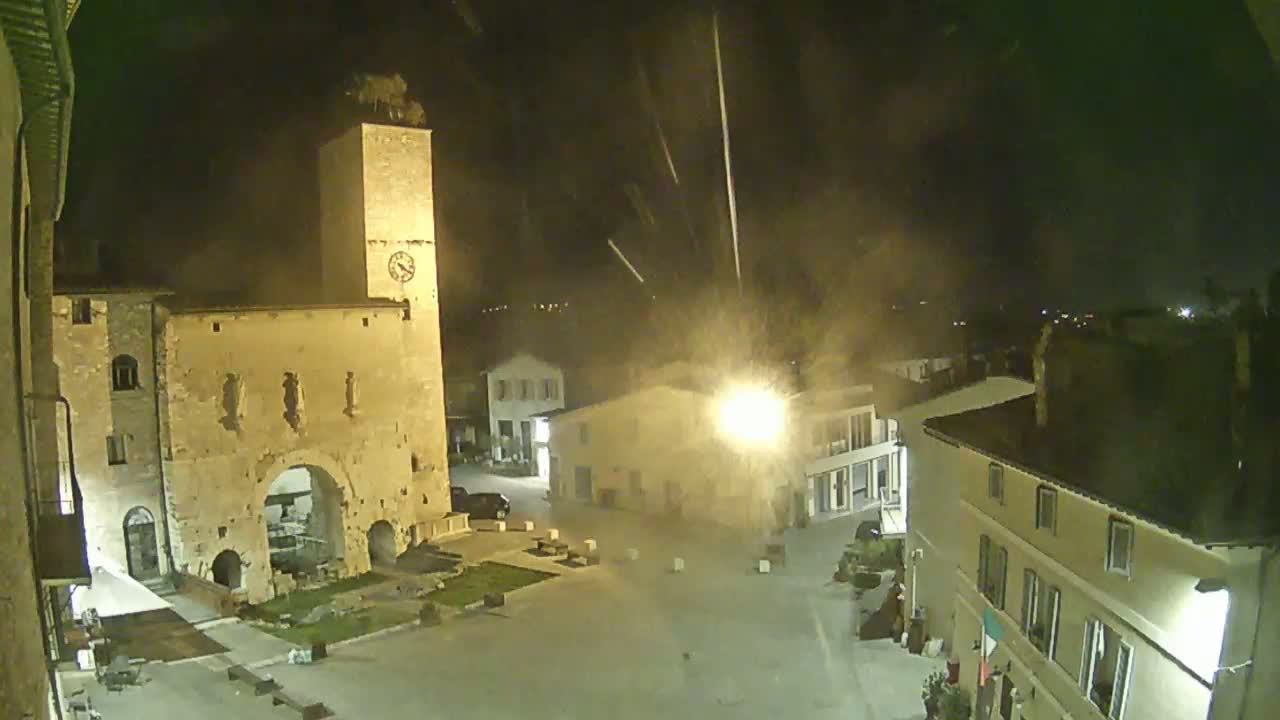 A nighttime view of a town square with a tall stone tower and several buildings, under a hazy sky with streaks of light possibly from rain or fireworks.