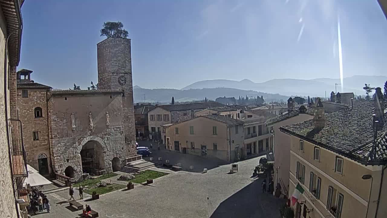 A sunny day reveals a town square surrounded by historic stone buildings, with a clock tower and distant hazy mountains in the background.