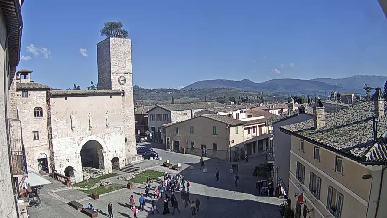 A sunny day reveals a town square in a historic Italian village, surrounded by stone buildings, with a clock tower and rolling hills in the background.