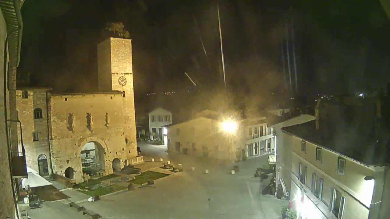 A nighttime, high-angle view of a town square with historic buildings, including a tall clock tower and archway, under a sky with streaks of light possibly from fireworks.