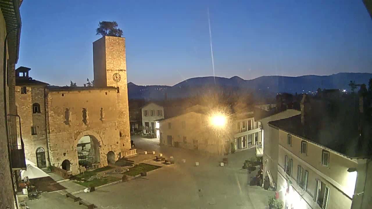 A town square at twilight is illuminated by streetlights, featuring a stone tower with a tree on top,  surrounded by older buildings against a backdrop of dark mountains.