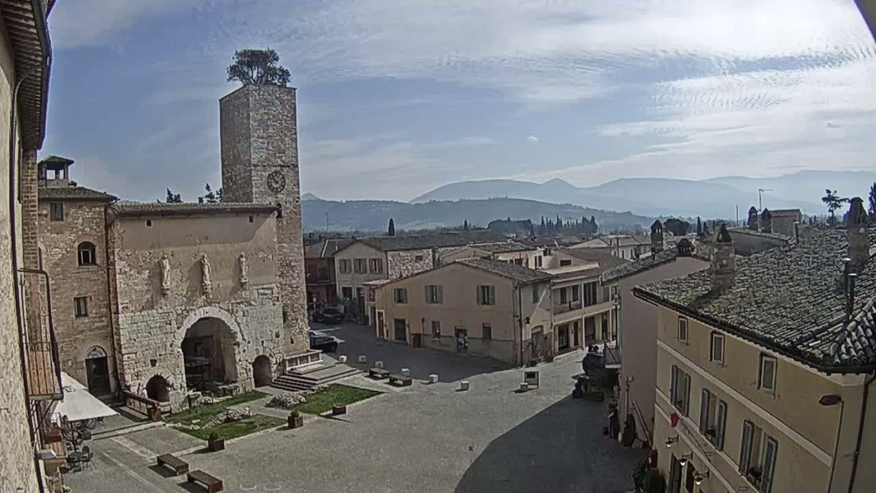 A town square in a historic Italian village, under a mostly clear sky, is surrounded by stone buildings, including a tall tower with a tree on top, and overlooks distant hazy mountains.