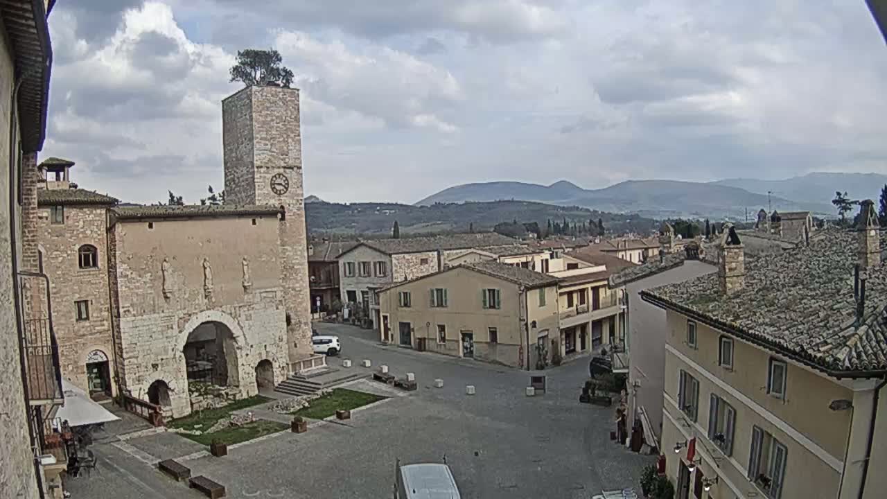 A town square in a small Italian town under a cloudy sky, overlooked by a tall stone tower with a tree on top, and mountains in the background.