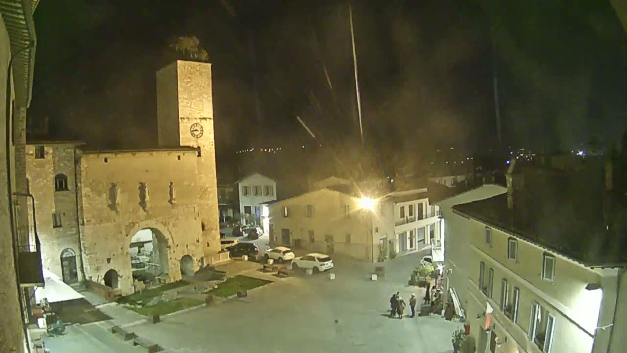 A nighttime view of a town square with several buildings, including a tall clock tower and a stone archway, under a slightly hazy sky with light streaks.