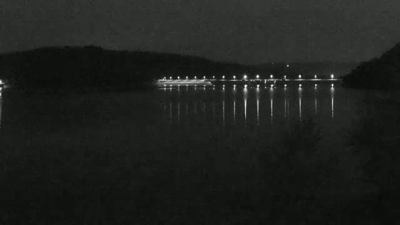 A nighttime view of a dark body of water reflecting the lights of a structure on the opposite shore, with a dark, tree-lined hill in the background.