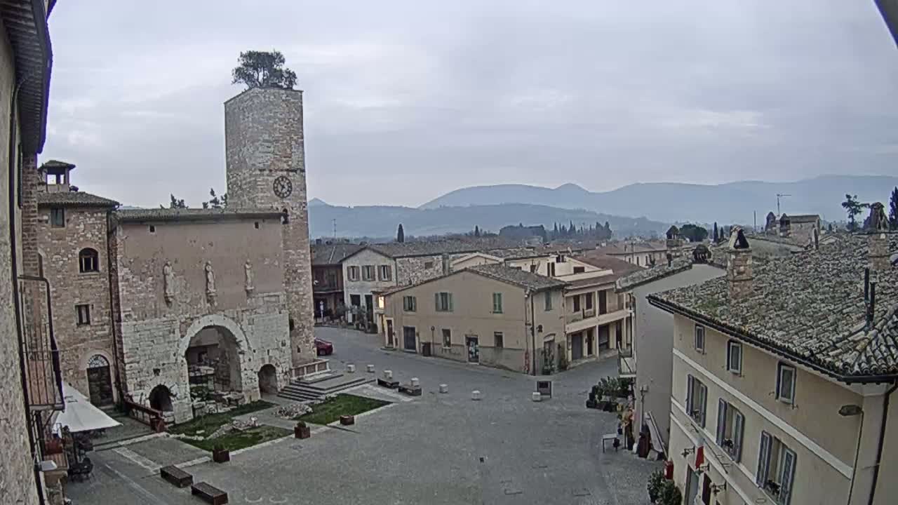 A town square in a historic Italian village, under an overcast sky, is surrounded by stone buildings, one with a clock tower topped with a tree.