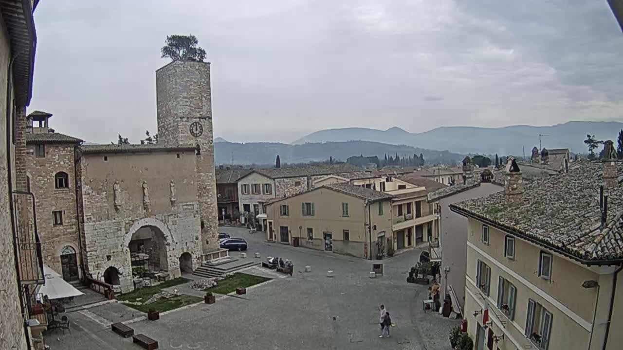 A town square in a medieval-style town, under an overcast sky, is surrounded by stone buildings, one featuring a tall clock tower with a tree on top, with distant mountains visible in the background.