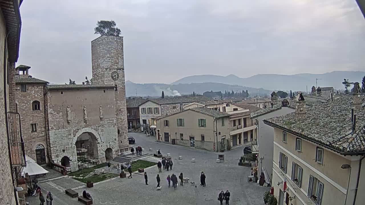 A town square in a medieval-style town, under an overcast sky, is populated by several people and dogs, with a tall brick tower and surrounding buildings visible.