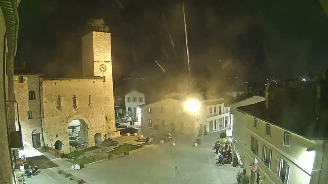 A nighttime view of a town square with a tall stone tower, several buildings, and a few people, under a dark sky with streaks of light.