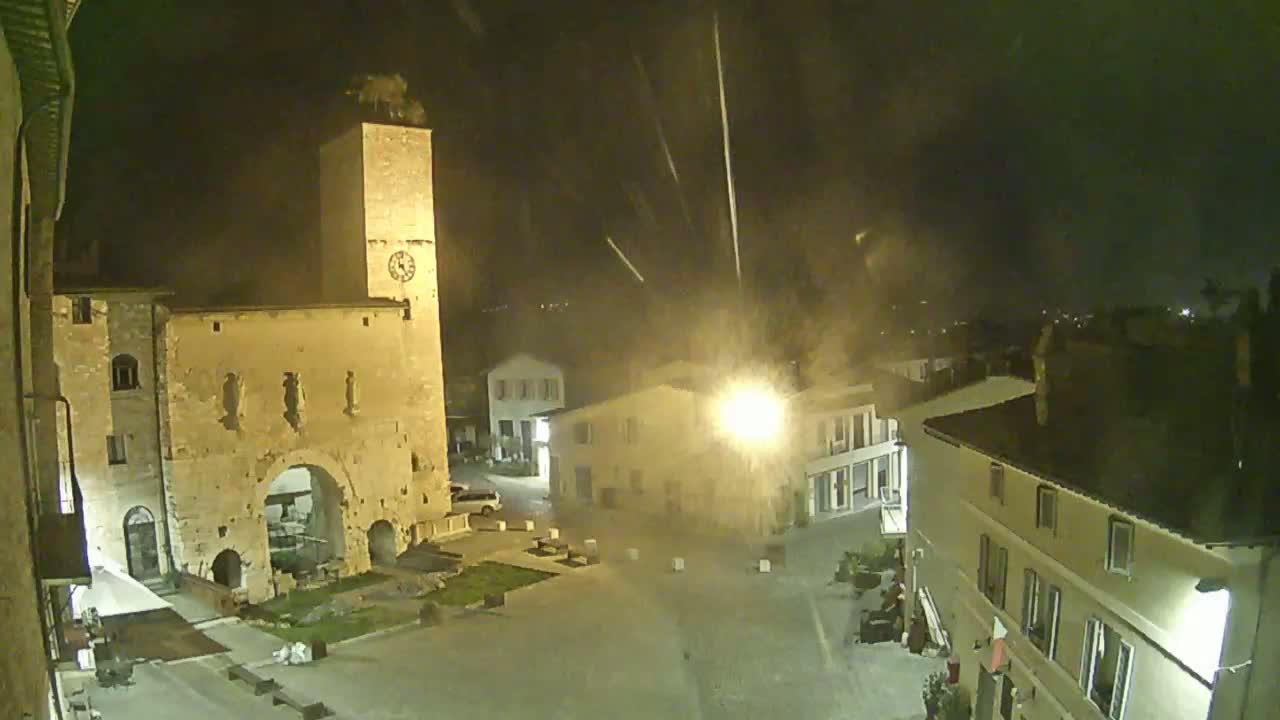 A nighttime view of a town square with old stone buildings, including a tall clock tower, under a lightly raining or snowing sky.