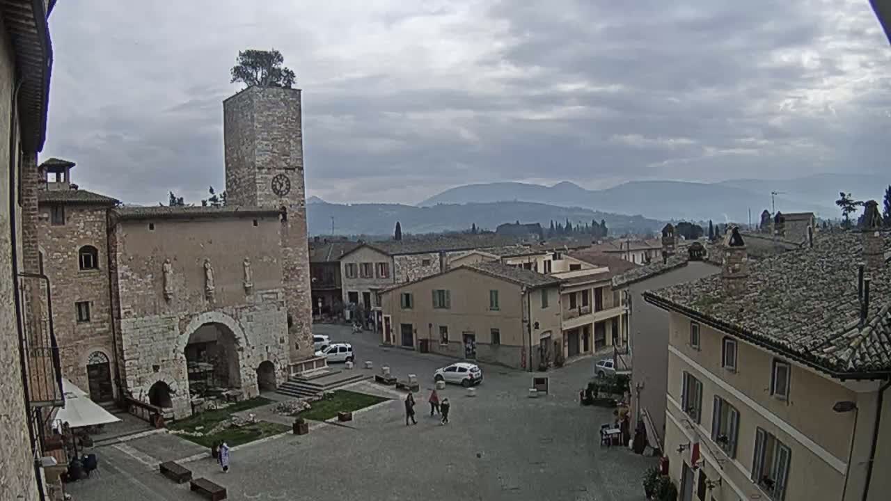 A stone town square with a tall clock tower, surrounded by buildings under an overcast sky, overlooks distant mountains.