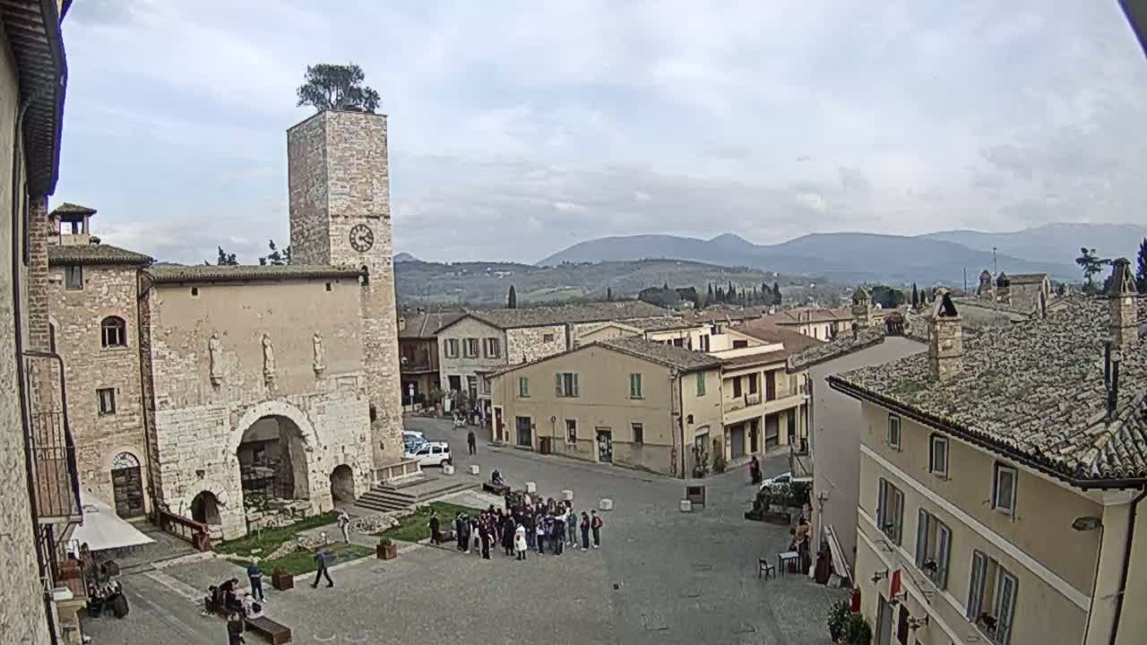 A town square in a hilltop town on a cloudy day, featuring a tall stone tower with a tree on top, several buildings, and a group of people gathered in the center.