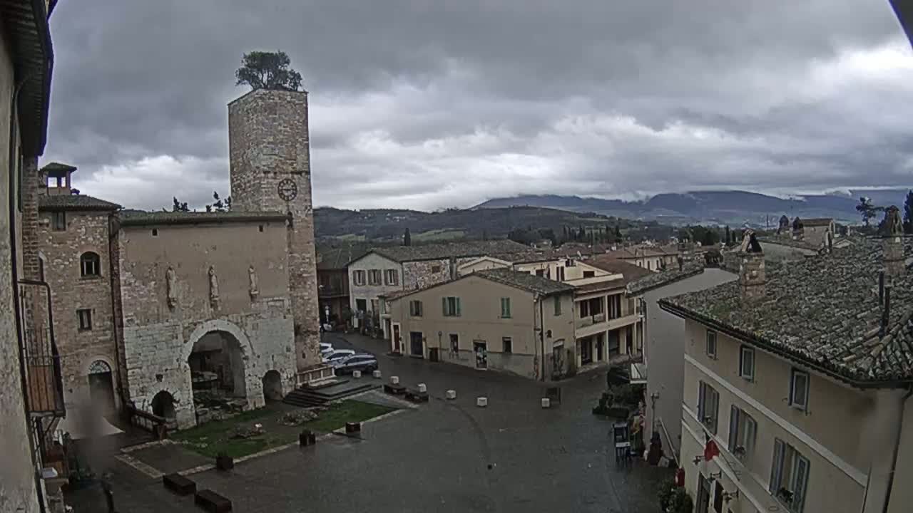 A town square in a historic Italian town, under an overcast sky, is surrounded by stone buildings, one featuring a tall tower with a tree on top.