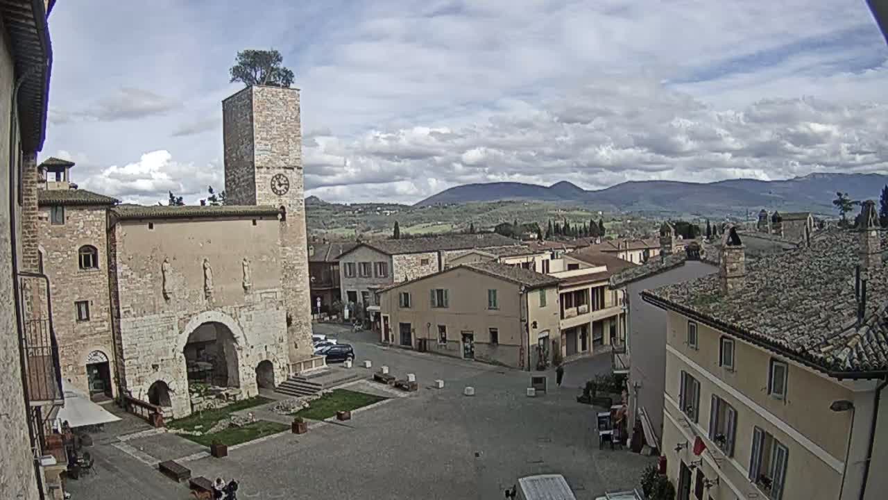 A town square in a historic Italian village under a partly cloudy sky, featuring a tall stone tower with a tree on top and surrounding buildings of similar architectural style.