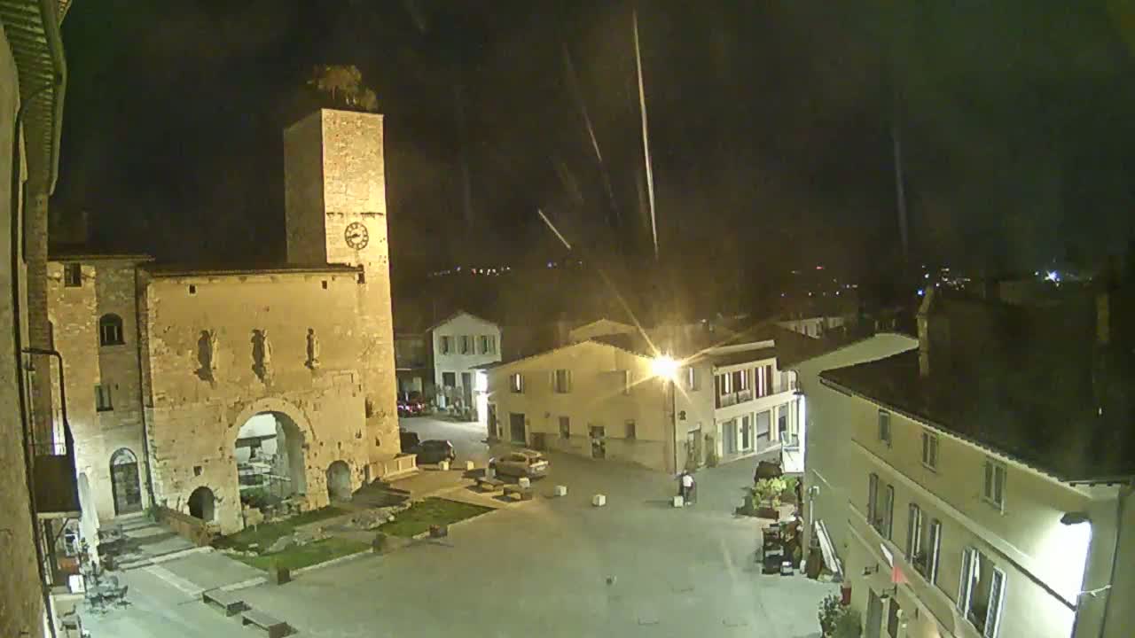 A nighttime view of a town square with several buildings, including a tall clock tower and a stone archway, under a lightly falling rain or snow.