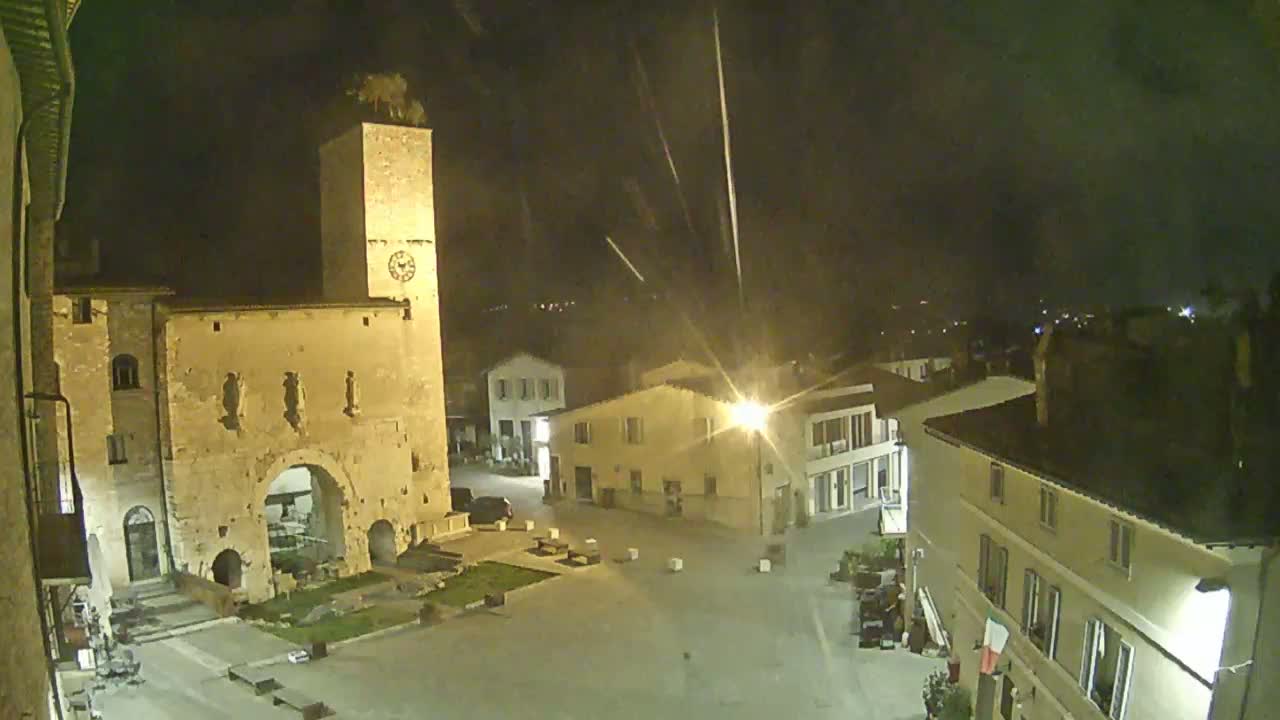 A nighttime view of a town square, illuminated by streetlights, with a tall clock tower and several buildings surrounding it under a partly cloudy sky.
