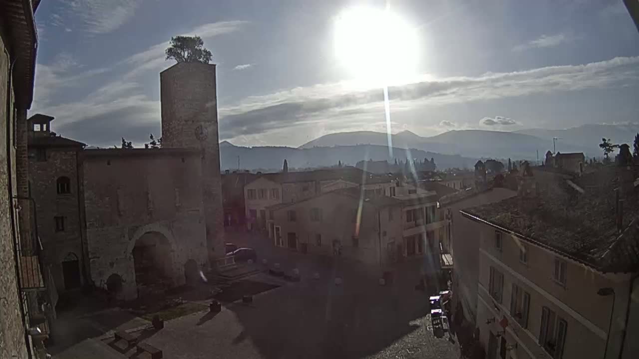 A sunlit view of a town square in a historic European town, with a tall tower topped with a tree, under mostly clear skies and mountains visible in the distance.