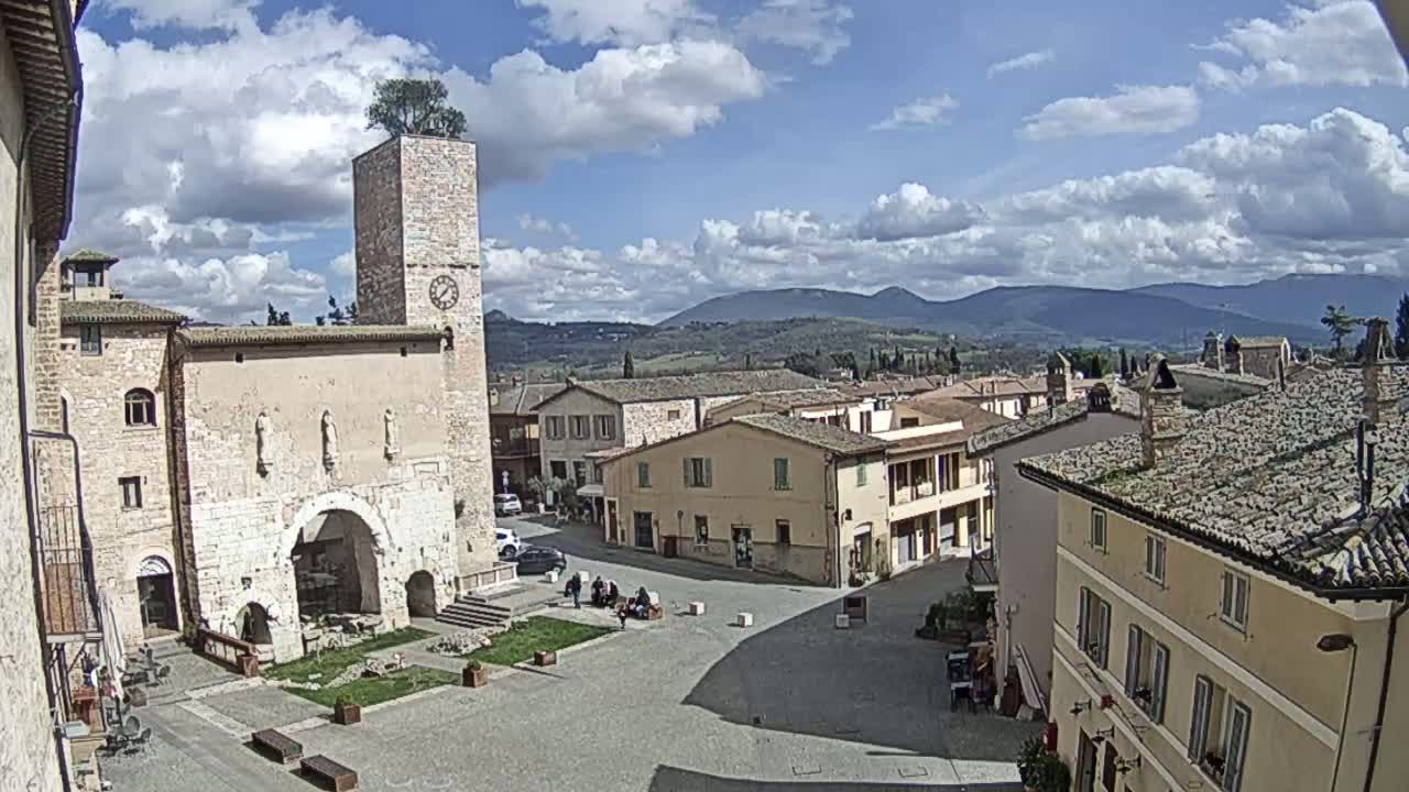 A town square in a village nestled in a valley under a partly cloudy sky, features a stone building with a clock tower and a tree on top.