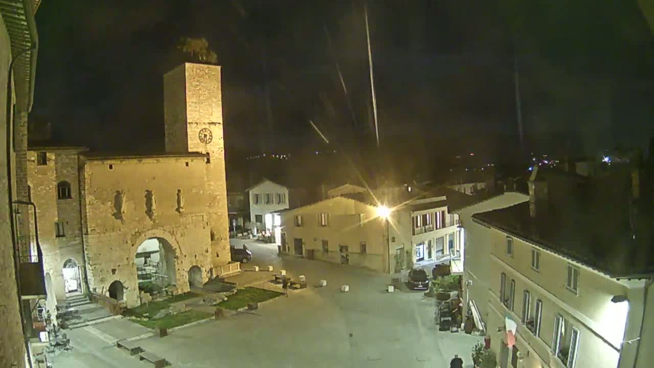 A nighttime view of a town square with old stone buildings, including a tall tower with a clock, under a dark sky with streaks of light.