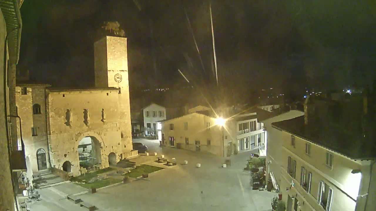 A nighttime view of a town square, featuring an ancient stone archway and clock tower, under a slightly rainy sky.