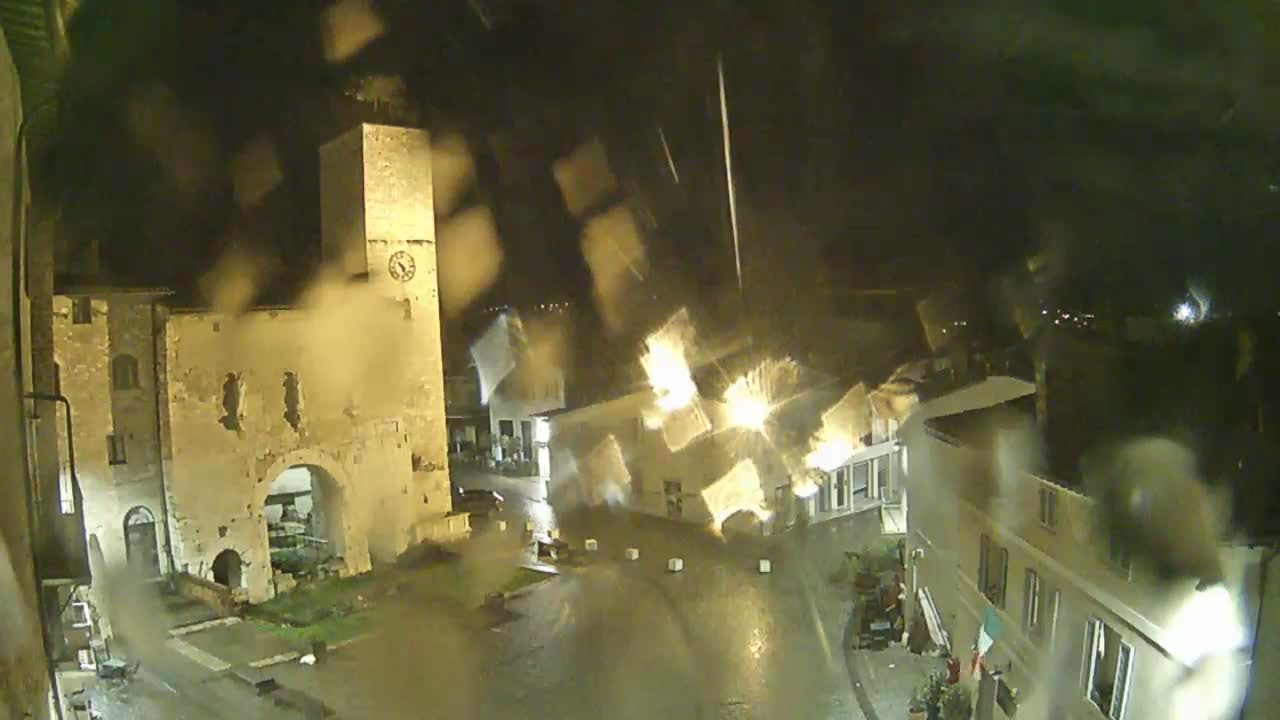 A rain-slicked town square at night, viewed through a rain-covered window, features an ancient stone building with a clock tower.