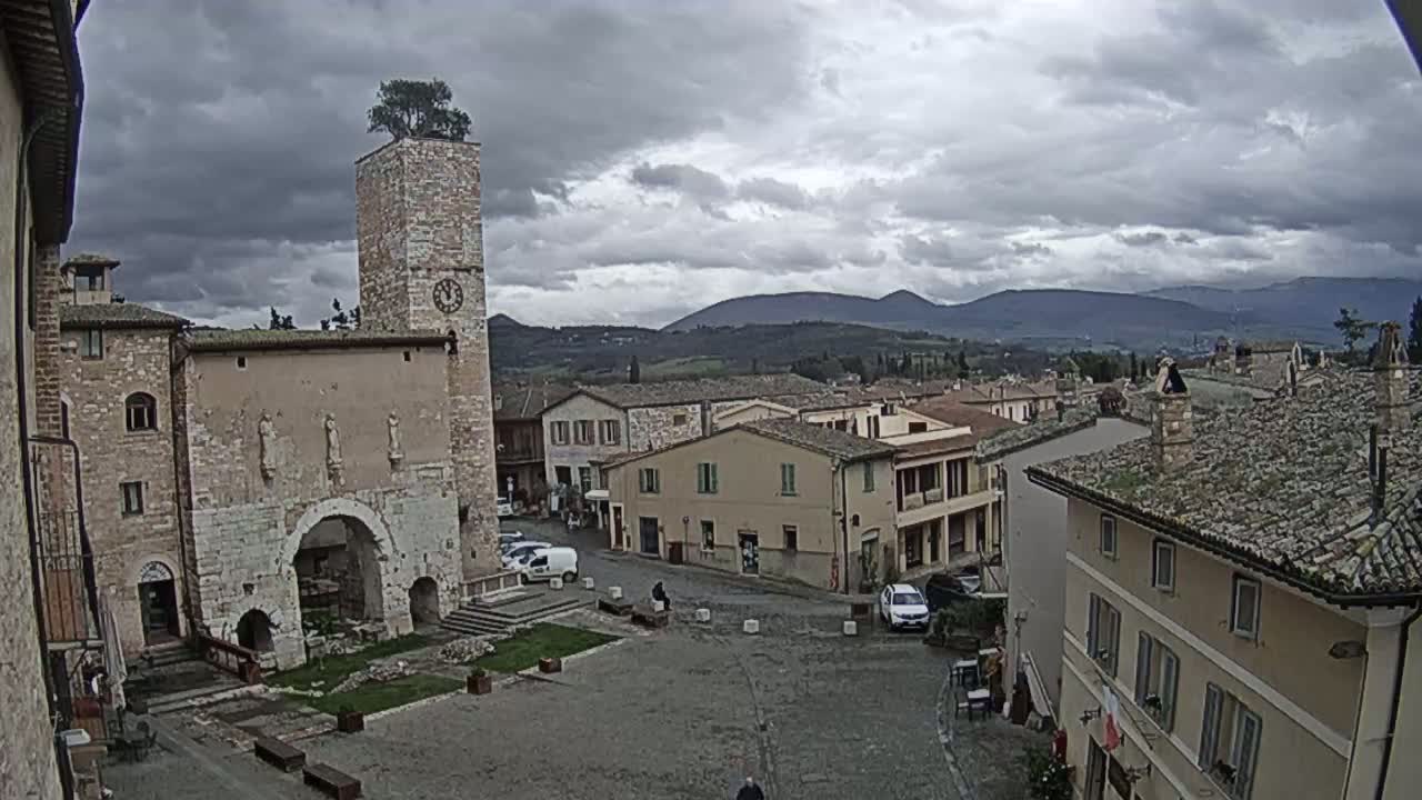 A town square in a village nestled in a valley, under a cloudy sky, features a stone building with a clock tower topped with a tree.