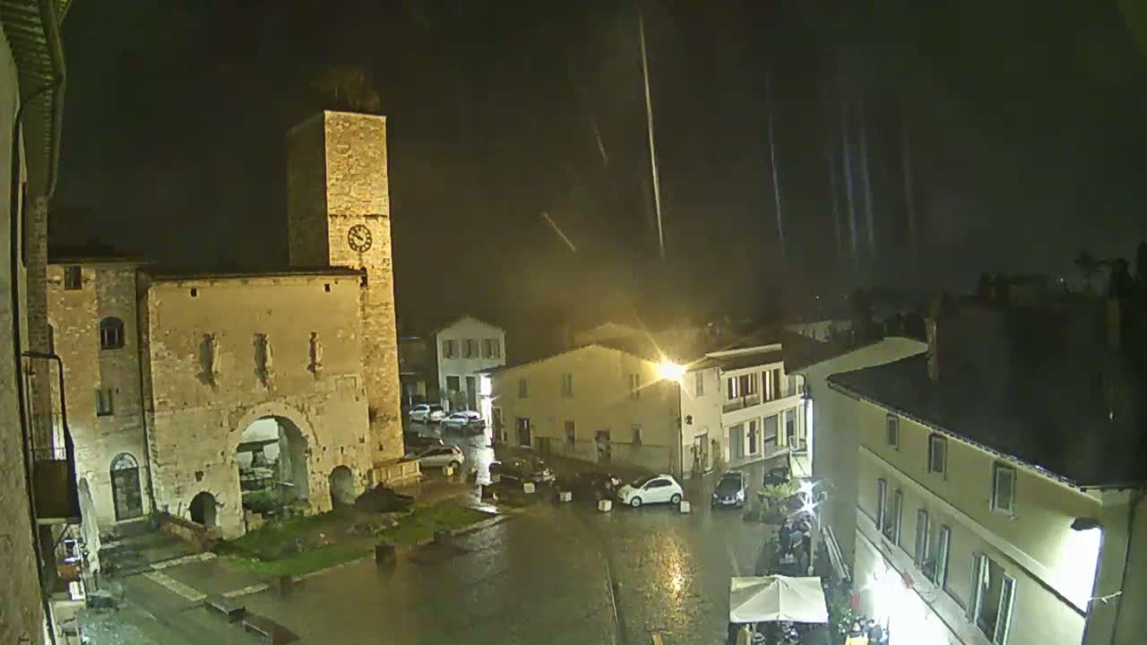 A rain-soaked town square at night, illuminated by streetlights, features a tall clock tower and several buildings.