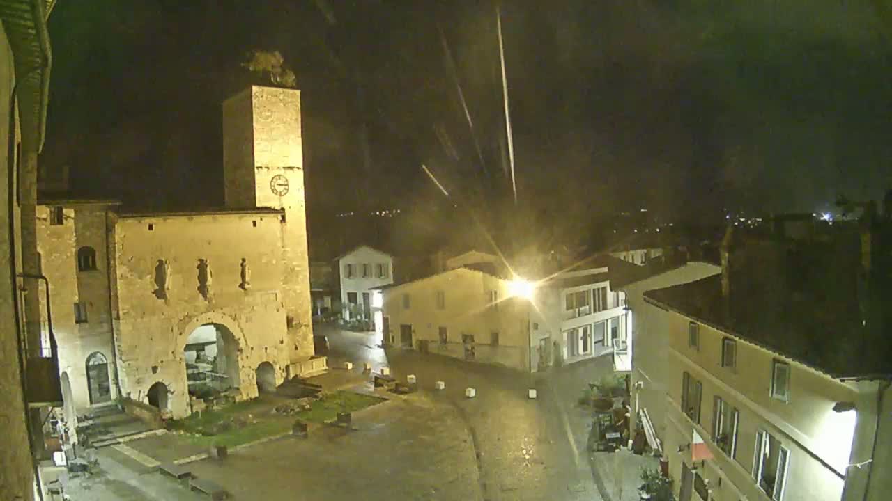 A nighttime view of a town square with a tall clock tower and several buildings under light rain.