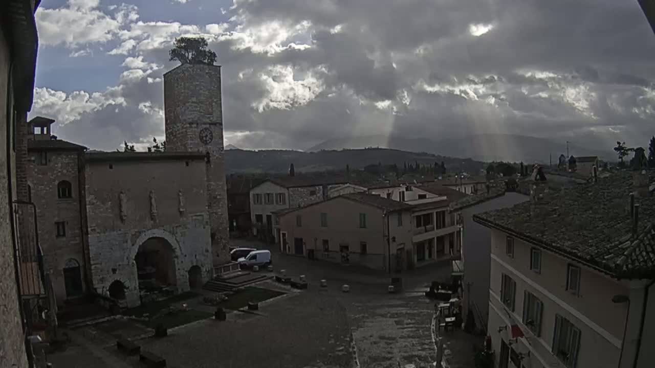 A town square with a stone tower and buildings under a cloudy sky with sunbeams breaking through.