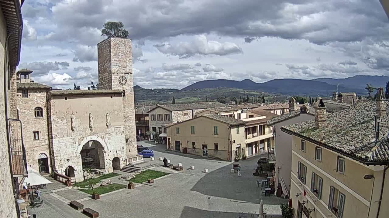 A town square in a village nestled in a valley, under a partly cloudy sky, features a stone building with a clock tower and a tree on top.