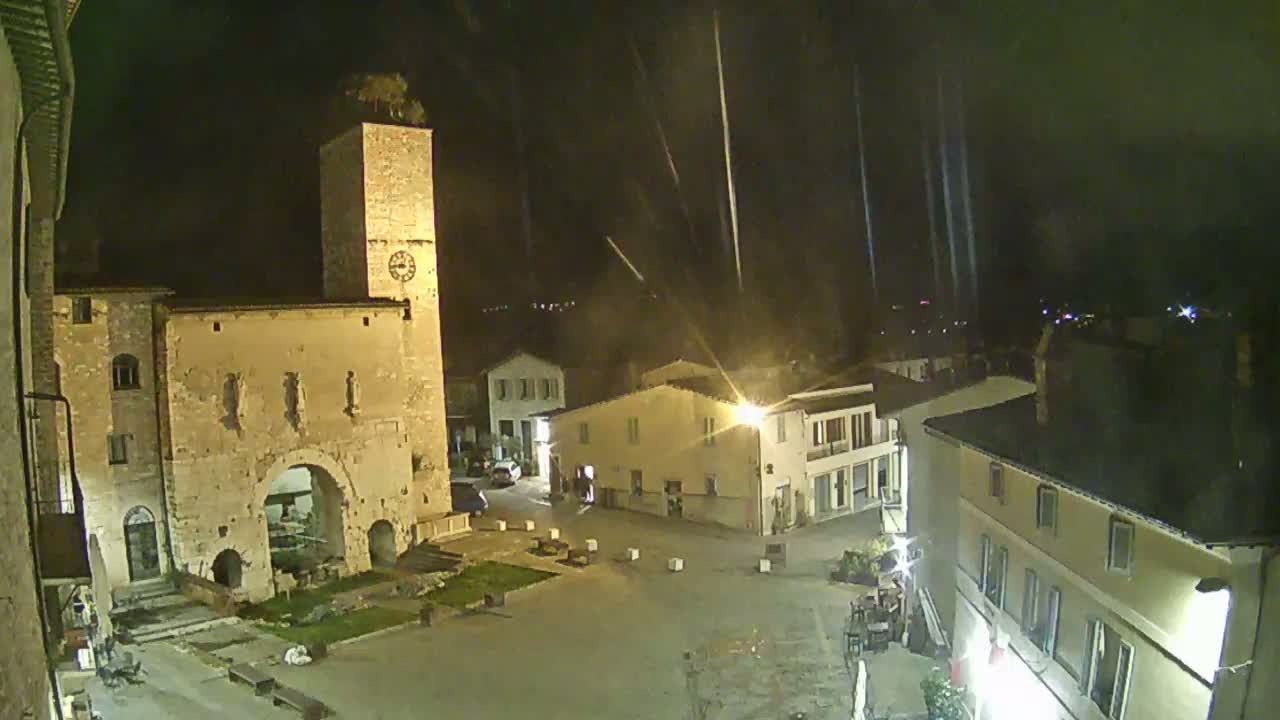 A nighttime view of a town square with old stone buildings, a tall clock tower, and light rain falling.
