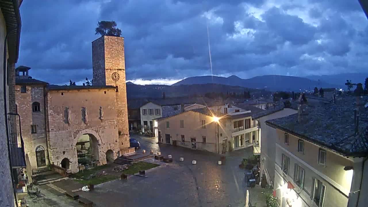 A town square at dusk, under a partly cloudy sky, features an ancient stone building with a clock tower, surrounding buildings, and distant mountains.