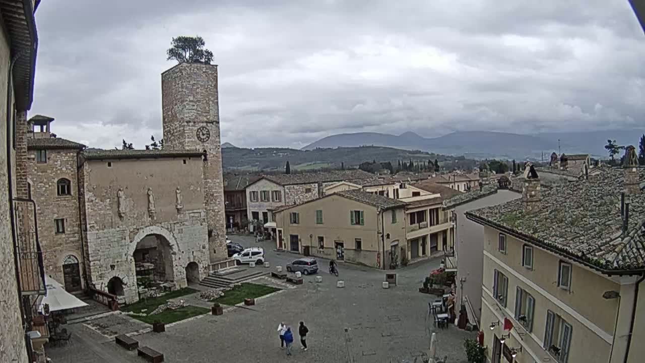 A town square with stone buildings, a clock tower topped with a tree, and distant mountains under a cloudy sky.