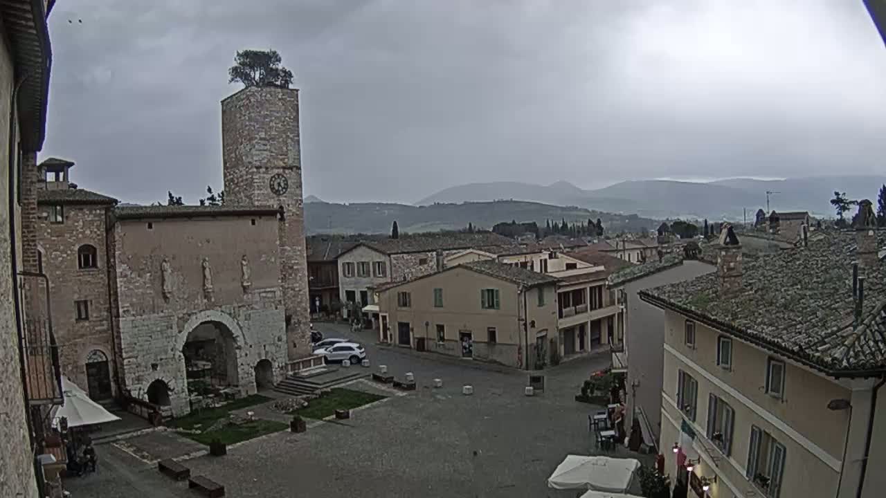 A town square in a hilltop village under an overcast sky, featuring a stone tower with a tree on top and several low buildings surrounding the square.
