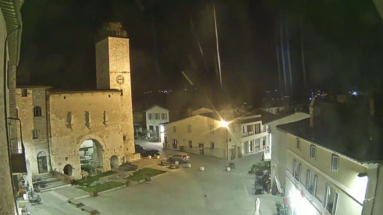 A nighttime view of a town square with stone buildings, a tall clock tower, and a few parked cars under a slightly rainy sky.