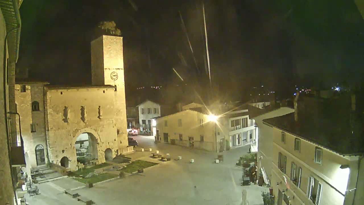 A nighttime view of a town square with a tall stone tower and several buildings under a slightly rainy sky.