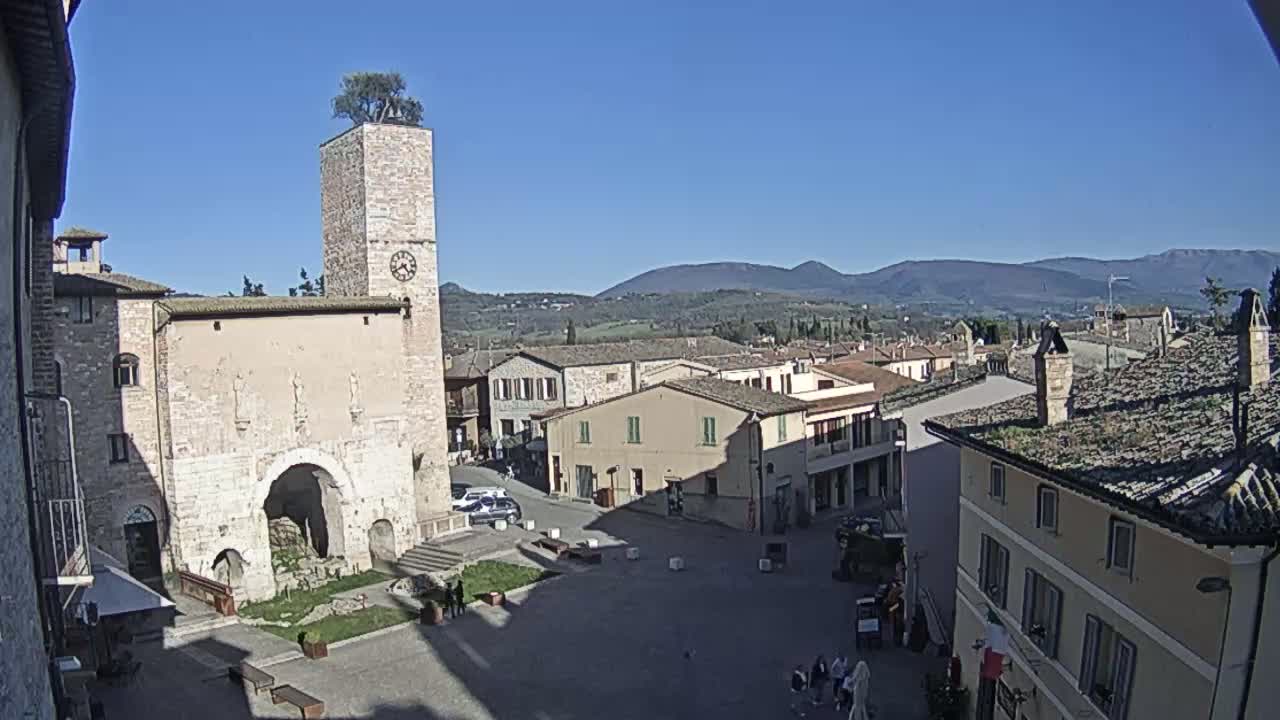 A town square in a small Italian town, under a clear blue sky, is surrounded by historic buildings and overlooks rolling hills in the distance.