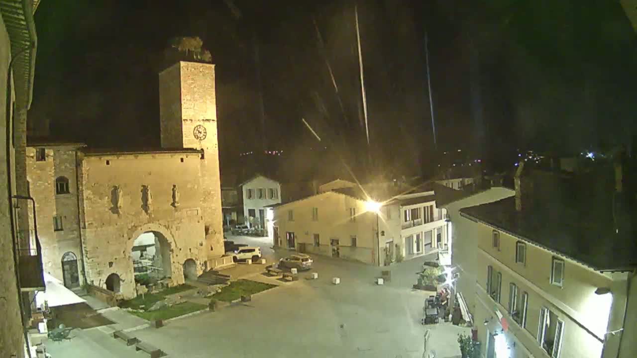 A nighttime view of a town square with a clock tower, several buildings, and a few cars, under a slightly rainy sky.