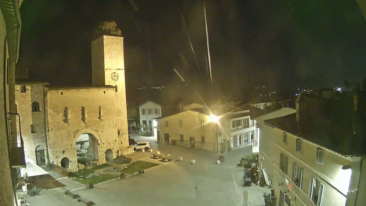 A nighttime view of a town square with a tall clock tower and several buildings, under a slightly hazy sky with light streaks.